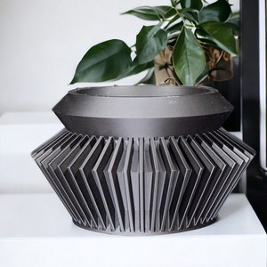 Peut inclure: Un pot de fleurs géométrique gris foncé avec un design angulaire unique. Le pot a une base large et cannelée et un dessus plus étroit, créant un contraste visuel saisissant. Le pot est posé sur une surface blanche, avec une plante verte en arrière-plan.