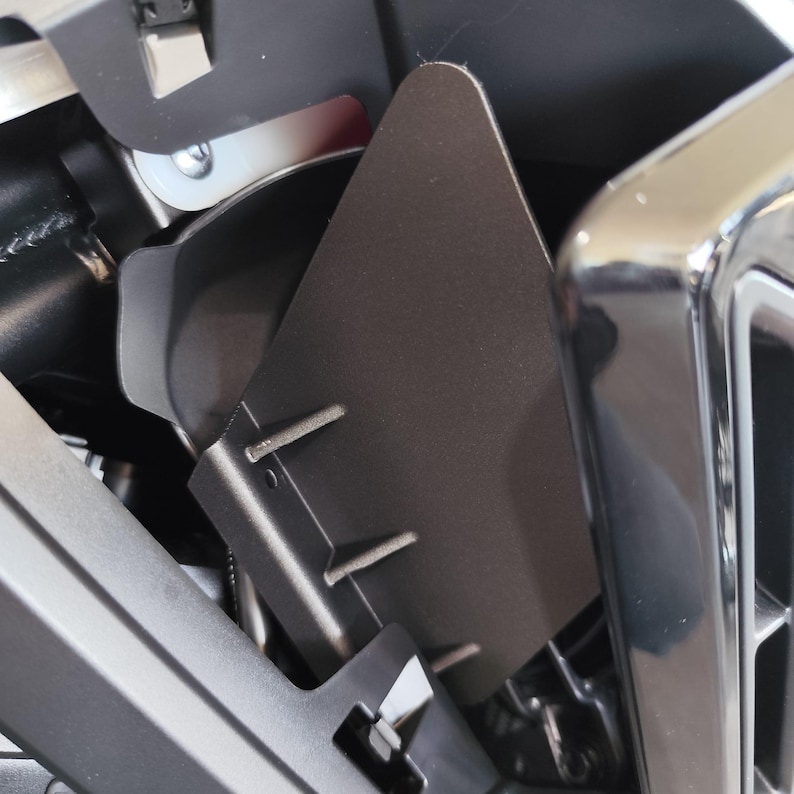 May include: Close-up of a dark gray metal component with a triangular shape and two small, parallel metal tabs. The component is part of a larger, complex assembly with other metal and plastic parts.
