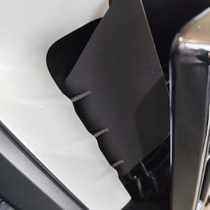 May include: Close-up of a black, angular automotive component with a textured surface. The component is positioned next to a white surface and a shiny, black metallic structure. The design features several small, gray protrusions.