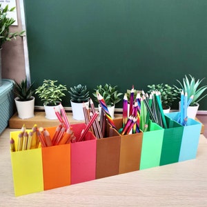 Può includere: Un set colorato di 10 porta matite in plastica stampata in 3D. I porta matite sono disposti a forma di arcobaleno, con ogni porta matite che contiene un colore diverso di matite. I porta matite sono seduti su una superficie di legno davanti a una lavagna verde.