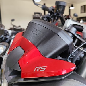 May include: Close-up of a red motorcycle fairing with a black plastic cover. The cover has the letters "RS" embossed on it. The fairing also has the letters "RS" in a silver colour on the red surface.
