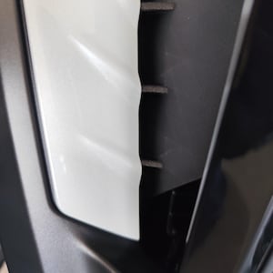 May include: Close-up of a section of a vehicle's interior. The image shows a white, textured panel next to a black, ridged component. The white panel has a pearlescent sheen, reflecting light. The black component has horizontal ridges.