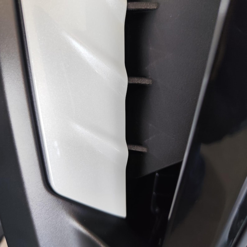 May include: Close-up of a section of a vehicle's interior. The image shows a white, textured panel next to a black, ridged component. The white panel has a pearlescent sheen, reflecting light. The black component has horizontal ridges.