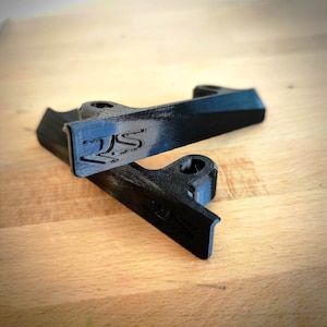 Peut inclure: Deux supports noirs imprimés en 3D avec une finition texturée. Chaque support présente un logo RS en relief sur la face avant et un trou de montage circulaire sur la bride supérieure. Les composants ont une forme angulaire et rigide.