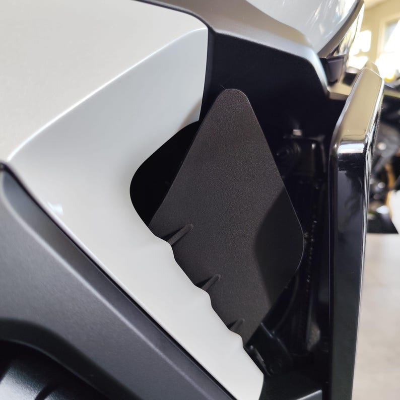 May include: Close-up of a motorcycle's front fairing, featuring a matte black, triangular insert. The insert is set against a backdrop of white and black body panels, with visible structural details. The design suggests a focus on aerodynamics and protection.