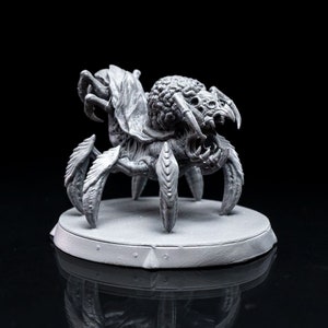 Op de afbeelding: Een grijs, gedetailleerd, 3D-geprint miniatuur van een spinachtig wezen met meerdere poten en een groot hoofd. Het wezen staat op een ronde basis.