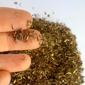 May include: A pile of gold metallic glitter, with a hand holding some of the glitter. The glitter is made of small, thin pieces of metal.