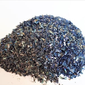 Könnte beinhalten: Ein Haufen Metallspäne mit einer Mischung aus blauen, silbernen und irisierenden Farben. Die kleinen, unregelmäßigen Stücke erzeugen eine strukturierte Optik, die das Licht reflektiert und einen schimmernden Effekt erzeugt. Das Material scheint industriell oder handwerklich zu sein.