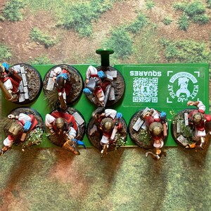 May include: Miniature figurines of fantasy characters, each mounted on a circular base with detailed terrain. The figures are dressed in red and white attire with blue accents. A green tray with the text "SQUARES" holds the bases.