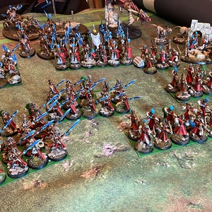 May include: A tabletop battle scene featuring miniature figures. The figures are arranged in formations on a green surface, with a variety of colors including red, blue, and white. The miniatures appear to be part of a fantasy or historical wargame.