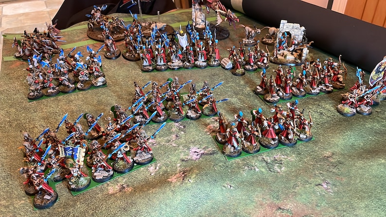 May include: A tabletop battle scene featuring miniature figures. The figures are arranged in formations on a green surface, with a variety of colors including red, blue, and white. The miniatures appear to be part of a fantasy or historical wargame.