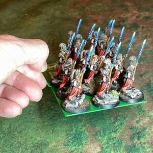 May include: A group of miniature fantasy figures on a green base. The figures are dressed in armor with red capes and wield blue-bladed spears. The bases are detailed with stone and grass, suggesting a battlefield setting.