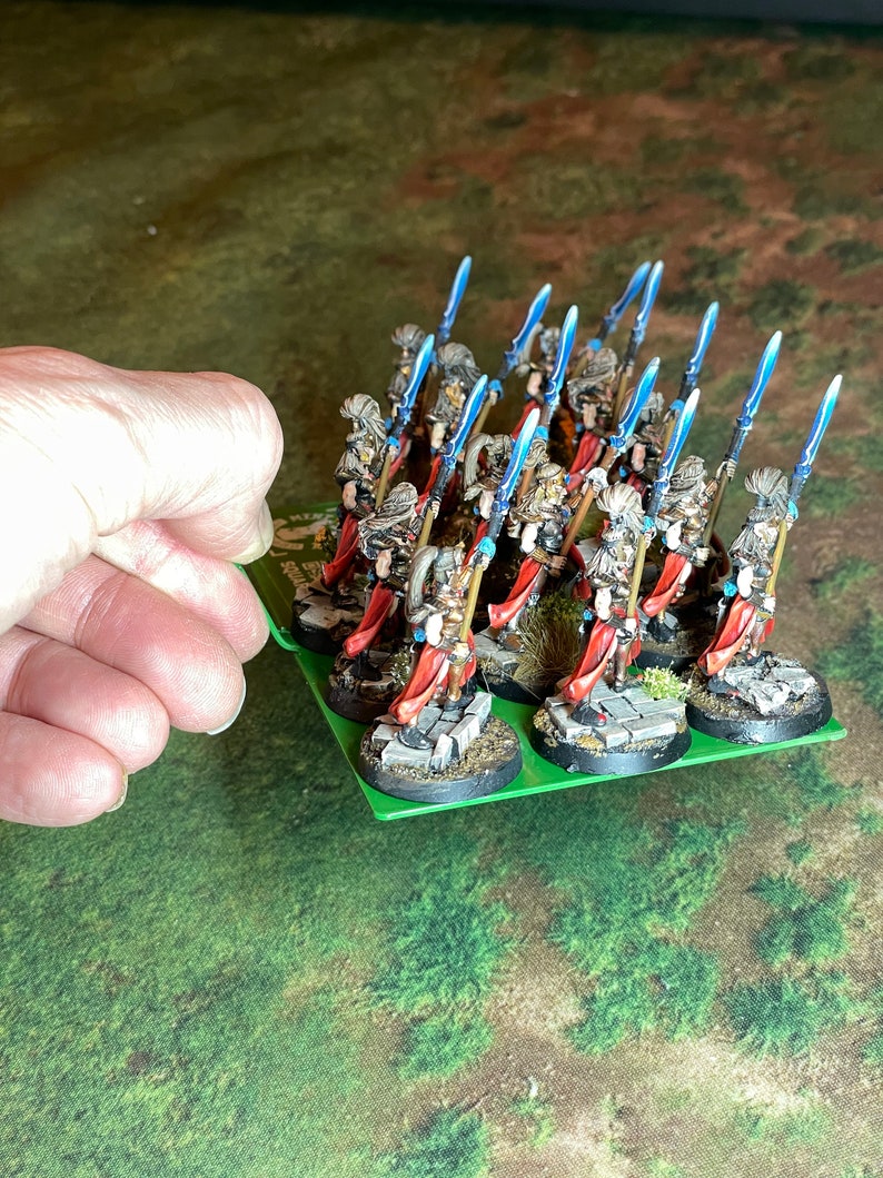 May include: A group of miniature fantasy figures on a green base. The figures are dressed in armor with red capes and wield blue-bladed spears. The bases are detailed with stone and grass, suggesting a battlefield setting.