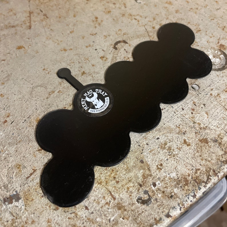 May include: Black metal tray with a unique cloud-like shape. The tray features a small handle and a circular sticker with the text "MINI MAG TRAY" and a graphic. The tray is designed to hold small items.