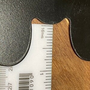 Puede incluir: Un objeto met&aacute;lico negro con forma curva, colocado junto a una regla blanca. La regla muestra medidas en dieciseisavos de pulgada. El objeto y la regla est&aacute;n sobre una superficie de madera.