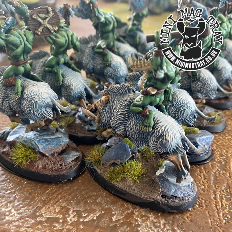 K&ouml;nnte beinhalten: Miniaturfiguren von gr&uuml;nen Orks, die auf grauen Wildschweinen reiten. Die Orks halten Waffen und Schilde. Die Wildschweine haben detailliertes Fell und Sto&szlig;z&auml;hne. Die Sockel sind mit Felsen und Gras verziert. Das Mini Mag Tray-Logo befindet sich oben rechts.