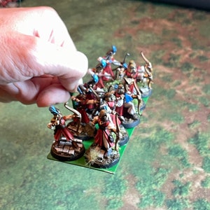 May include: Miniature figurines of archers in red and white robes with blue accents, holding bows. The figurines are on a green base, held by a hand. The background is a green and brown surface.