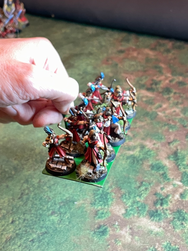 May include: Miniature figurines of archers in red and white robes with blue accents, holding bows. The figurines are on a green base, held by a hand. The background is a green and brown surface.