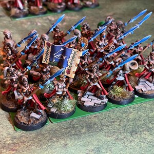 May include: Miniature fantasy figures of warriors with spears and a banner. The figures are painted with gold, red, and blue accents. The banner is blue with a white emblem. The figures are on round bases with miniature foliage.