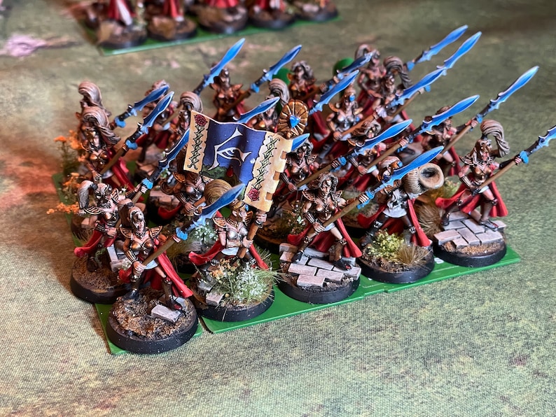 May include: Miniature fantasy figures of warriors with spears and a banner. The figures are painted with gold, red, and blue accents. The banner is blue with a white emblem. The figures are on round bases with miniature foliage.
