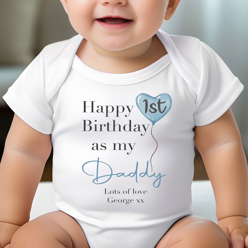 Pode incluir: Um macac&atilde;o branco para beb&ecirc; com texto azul que diz "Feliz 1&ordm; anivers&aacute;rio como meu papai. Muito amor George xx". Um bal&atilde;o azul em forma de cora&ccedil;&atilde;o com o n&uacute;mero "1&ordm;" est&aacute; acima do texto.