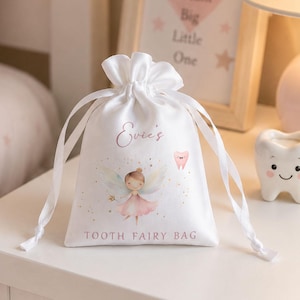 May include: A white satin drawstring pouch featuring the text Evie's and TOOTH FAIRY BAG in pink lettering. The bag displays a graphic of a fairy with wings and a pink dress, accompanied by a small pink tooth illustration and gold sparkles.