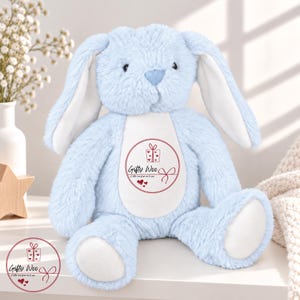 May include: A light blue plush rabbit with long ears, a white belly patch, and white foot pads. The belly features a circular red graphic with a gift box icon, the text Gifty Woo, and the phrase A little love from me to you with hearts.