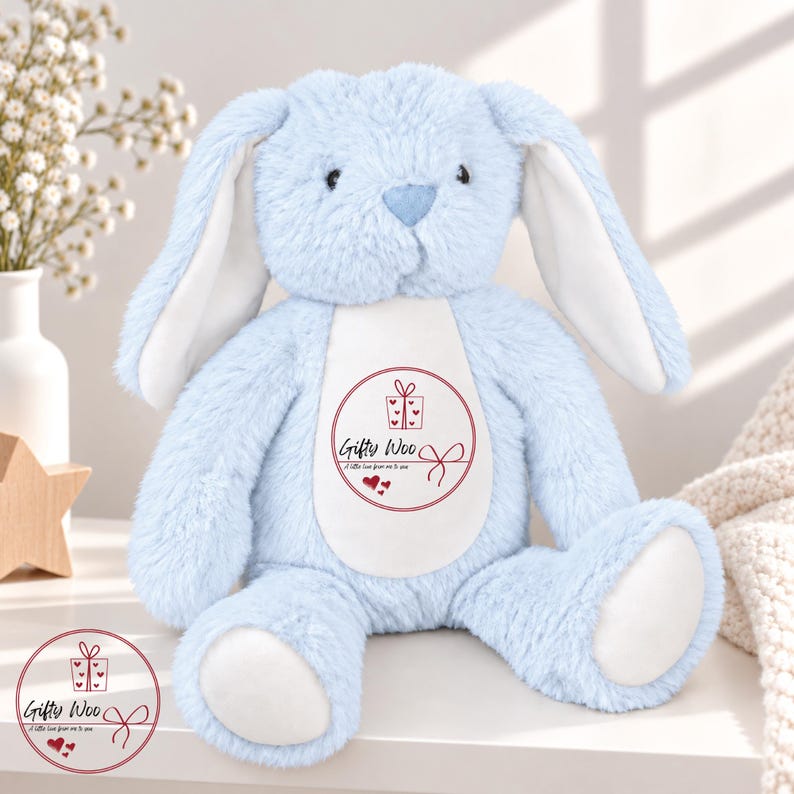 May include: A light blue plush rabbit with long ears, a white belly patch, and white foot pads. The belly features a circular red graphic with a gift box icon, the text Gifty Woo, and the phrase A little love from me to you with hearts.
