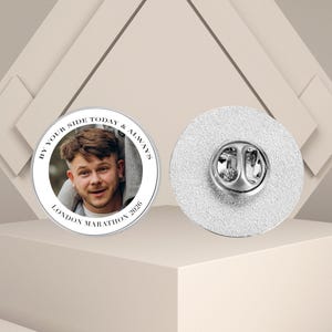 May include: A circular silver pin featuring a photograph of a person, with the text "BY YOUR SIDE TODAY & ALWAYS" and "LONDON MARATHON 2026" encircling the image. The pin has a silver clasp on the reverse.