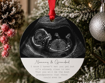 Può includere: Un ornamento natalizio rotondo e piatto con un'ecografia fetale in bianco e nero. Sotto l'immagine, il testo recita: Nanny & Grandad, I know I haven't met you yet but it's already clear to see That you will be the best grandparents that there could ever be. Un nastro rosso è fissato alla parte superiore.