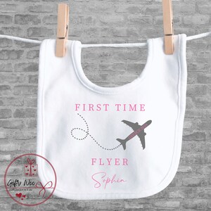 Może przedstawiać: Biały śliniaczek dla niemowlęcia z r&oacute;żowym nadrukiem samolotu i tekstem "First Time Flyer Sophia" w kolorze r&oacute;żowym.