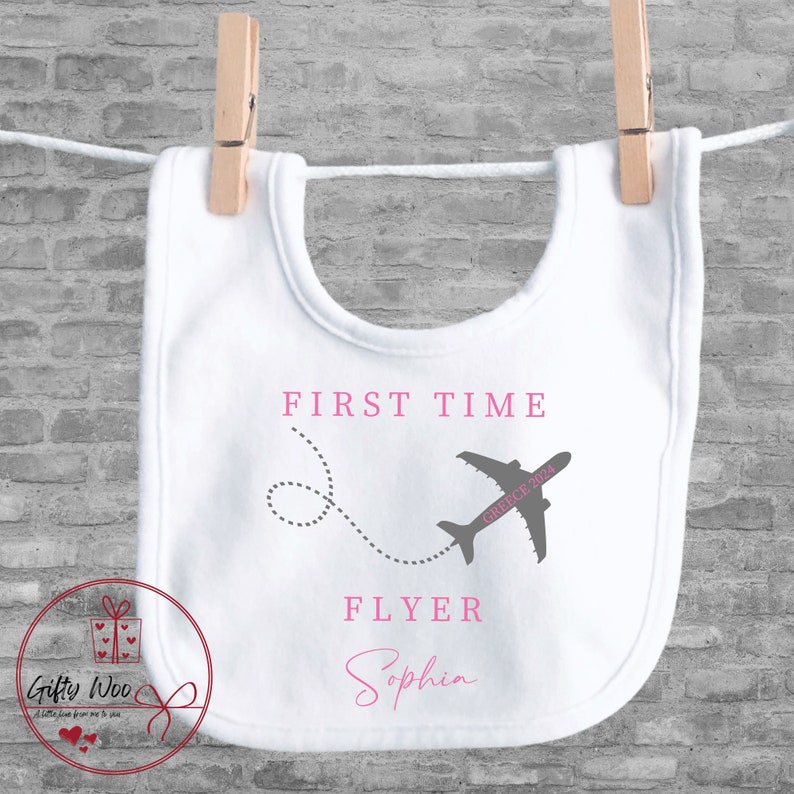 Może przedstawiać: Biały śliniaczek dla niemowlęcia z r&oacute;żowym nadrukiem samolotu i tekstem "First Time Flyer Sophia" w kolorze r&oacute;żowym.