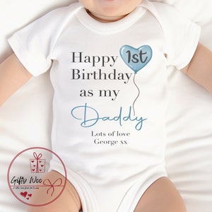 Pode incluir: Um macac&atilde;o branco para beb&ecirc; com texto azul que diz "Feliz 1&ordm; anivers&aacute;rio como meu papai" com um bal&atilde;o azul em forma de cora&ccedil;&atilde;o. O texto "Muito amor George xx" tamb&eacute;m est&aacute; inclu&iacute;do.