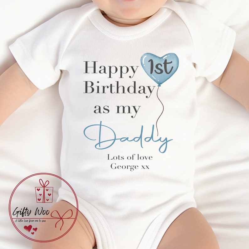 Pode incluir: Um macac&atilde;o branco para beb&ecirc; com texto azul que diz "Feliz 1&ordm; anivers&aacute;rio como meu papai" com um bal&atilde;o azul em forma de cora&ccedil;&atilde;o. O texto "Muito amor George xx" tamb&eacute;m est&aacute; inclu&iacute;do.