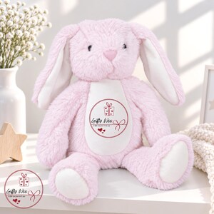 May include: A plush pink rabbit toy with long ears, a white belly patch, and white foot pads. The belly features a circular graphic with a gift box icon, three small hearts, and the text Gifty Woo A little love from me to you.