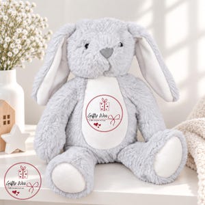 May include: A plush grey rabbit toy with long ears and a white belly patch. The belly features a circular red graphic with a gift box icon, the text "Gifty Woo", the phrase "A little love from me to you", and three small red hearts.