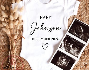 Pode incluir: Body de bebé branco com o texto "BABY Johnson DECEMBER 2026" e um símbolo de coração. Três ecografias são colocadas ao lado do body. O body está sobre uma superfície tecida com flores secas.