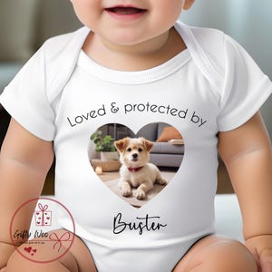 May include: White baby onesie with a heart-shaped image of a puppy inside. The text "Loved & protected by" is above the image and the name "Buster" is below the image.
