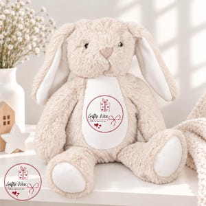 May include: A beige plush bunny toy with long floppy ears and a white belly patch. The belly features a circular red graphic with a gift box icon, the text "Gifty Woo", the phrase "A little love from me to you", and three small red hearts.