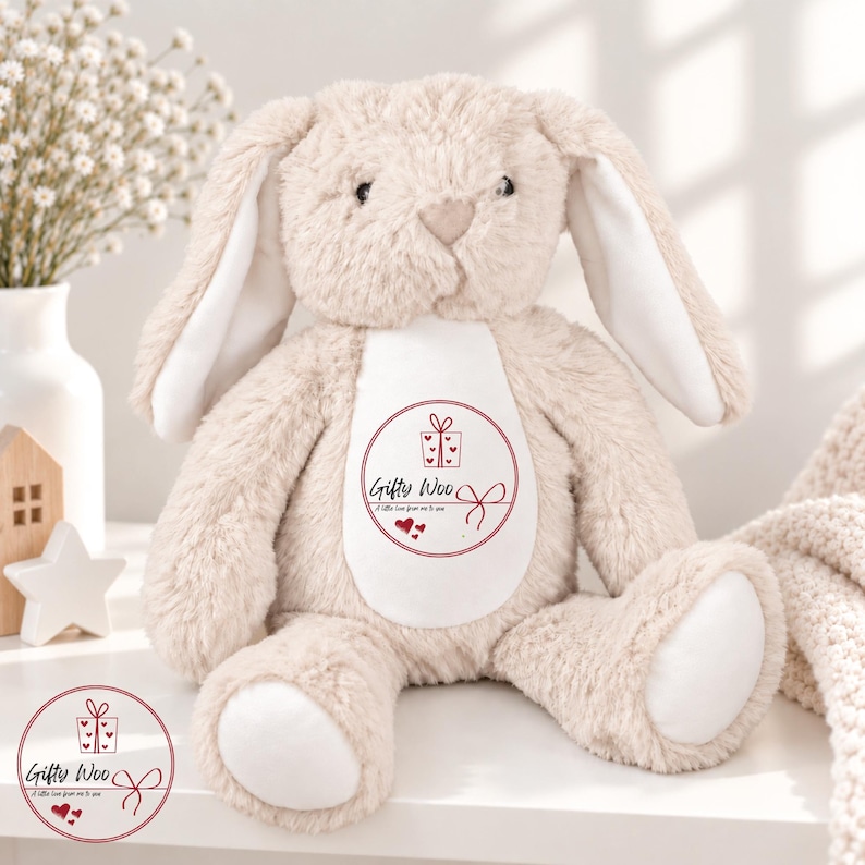 May include: A beige plush bunny toy with long floppy ears and a white belly patch. The belly features a circular red graphic with a gift box icon, the text "Gifty Woo", the phrase "A little love from me to you", and three small red hearts.