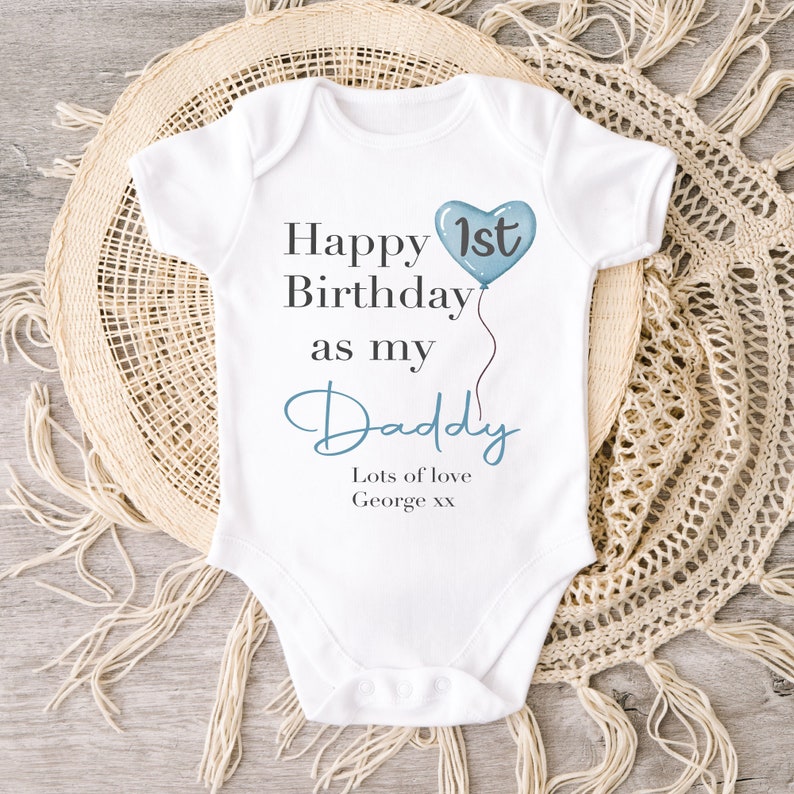 Pode incluir: Macac&atilde;o branco para beb&ecirc; com um bal&atilde;o em forma de cora&ccedil;&atilde;o azul que diz "1&ordm;" e o texto "Feliz anivers&aacute;rio como meu papai. Muito amor George xx".