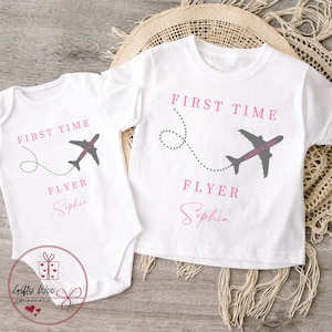 Może przedstawiać: Dwie białe koszulki z napisem "First Time Flyer" i grafiką szarego samolotu. Mniejsza koszulka ma imię "Sophia" napisane r&oacute;żowym kolorem. Większa koszulka ma imię "Sophia" napisane r&oacute;żowym kolorem i napis "Grecja 2024" na samolocie.