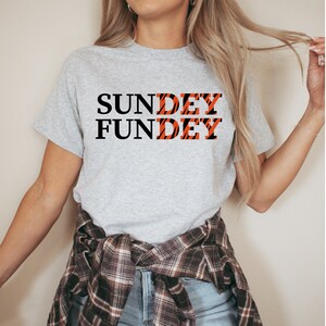 May include: A heather gray short-sleeve t-shirt featuring the black text "SUNDAY FUNDAY" printed on the front. The letters "DEY" in both words are filled with an orange and black tiger stripe pattern. A plaid flannel shirt is tied around the waist.