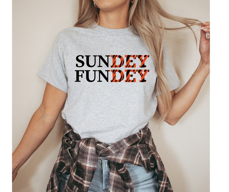 May include: A heather gray short-sleeve t-shirt featuring the black text "SUNDAY FUNDAY" printed on the front. The letters "DEY" in both words are filled with an orange and black tiger stripe pattern. A plaid flannel shirt is tied around the waist.