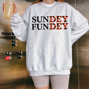 May include: A light gray crewneck sweatshirt featuring the black text "SUNDEY" and "FUNDEY" printed on the front. The letters "DEY" in both words are filled with an orange and black tiger stripe pattern. The sweatshirt has long sleeves and a ribbed hem.