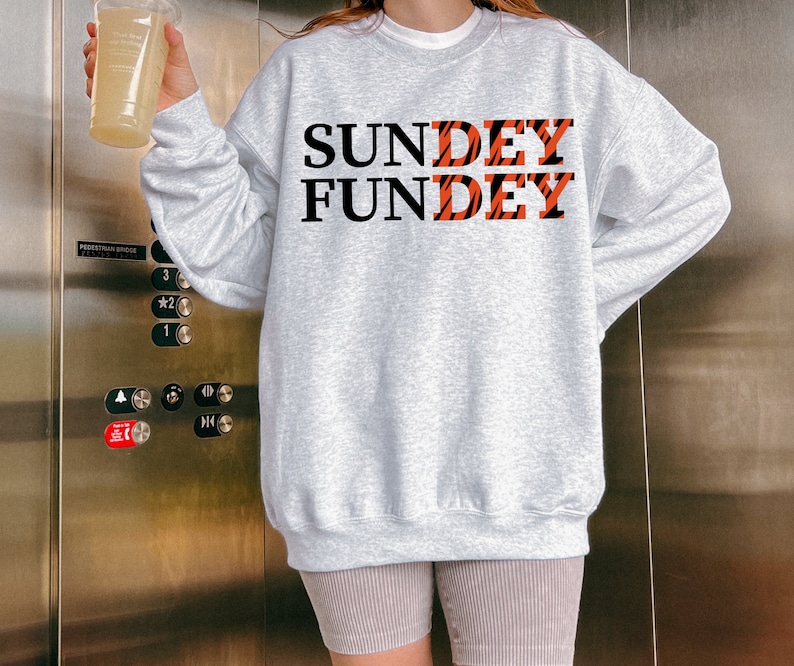 May include: A light gray crewneck sweatshirt featuring the black text "SUNDEY" and "FUNDEY" printed on the front. The letters "DEY" in both words are filled with an orange and black tiger stripe pattern. The sweatshirt has long sleeves and a ribbed hem.