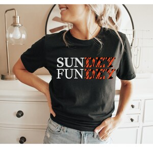 May include: A black short-sleeved t-shirt featuring the white text "SUN" and "FUN" stacked vertically on the left, with the word "DAY" appearing twice in a black and orange tiger-stripe pattern to the right of each word, creating the phrase "SUNDAY FUNDAY".