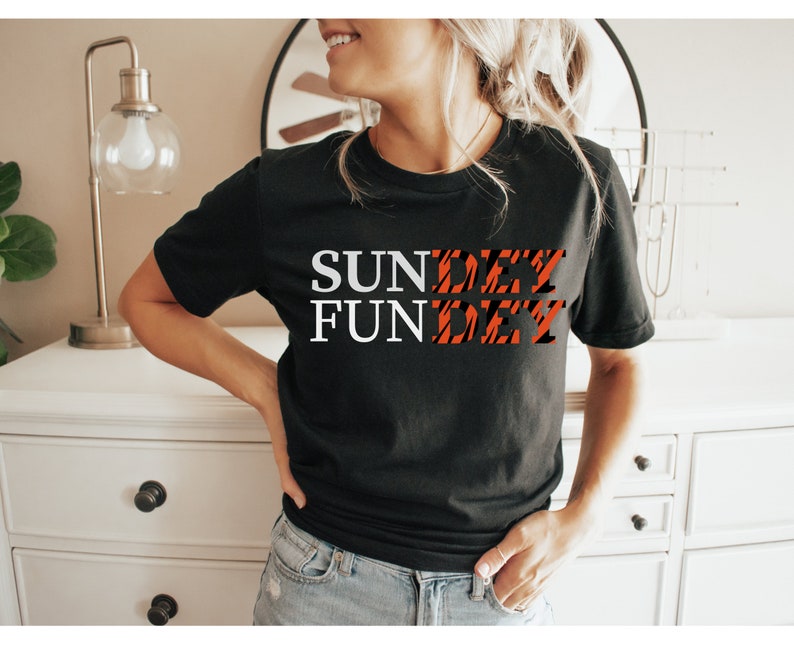 May include: A black short-sleeved t-shirt featuring the white text "SUN" and "FUN" stacked vertically on the left, with the word "DAY" appearing twice in a black and orange tiger-stripe pattern to the right of each word, creating the phrase "SUNDAY FUNDAY".
