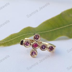 Puede incluir: Un anillo dorado con un diseño en forma de cruz. El anillo presenta una gema roja central rodeada de seis gemas rosas redondas más pequeñas. El anillo está sobre un fondo blanco con una hoja verde.