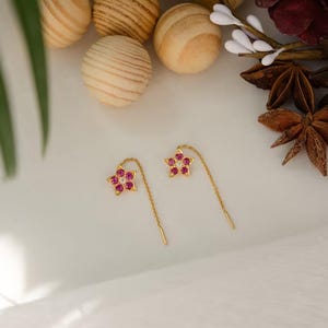 May include: A pair of gold star-shaped earrings, each with red gemstones and a central white gemstone. The earrings feature a fine chain. The earrings are displayed on a white surface.